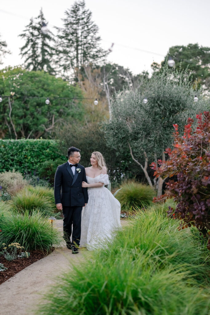 Melanie & Hang plan a backyard wedding at a private residence. Photo of couple on wedding day walking through the property greenery with string lights. 