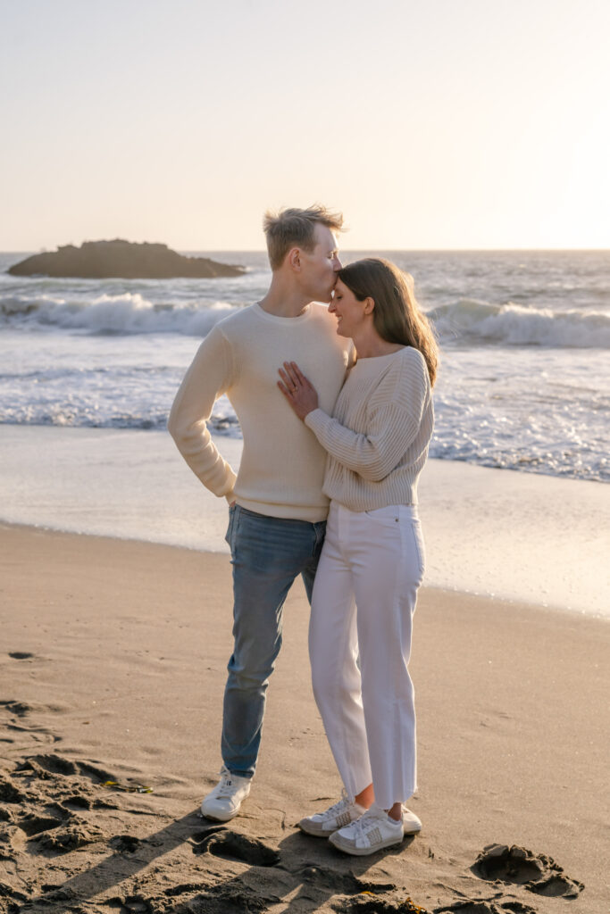 Engaged couple standing together on Baker Beach in San Francisco during their proposal session, with ocean waves and soft evening light in the background.