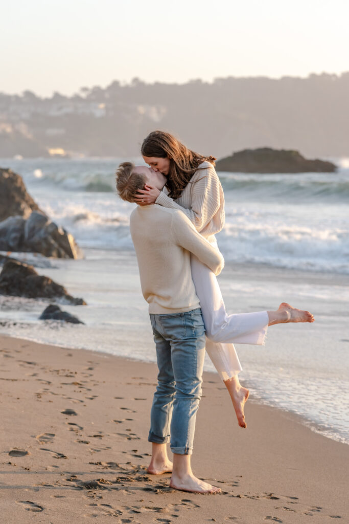 Newly engaged couple sharing a joyful embrace and kiss at Baker Beach in San Francisco following their proposal, photographed along the shoreline at sunset.