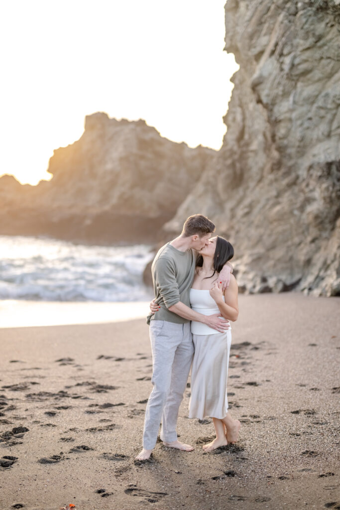 Newly engaged couple embracing and kissing on the beach near Coit Tower in San Francisco following their proposal, with rocky cliffs and ocean waves behind them.