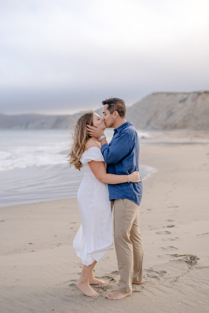 Newly engaged couple kissing on the beach near the Marin Headlands after their proposal, with the coastline and cliffs in the background in Marin County, California.