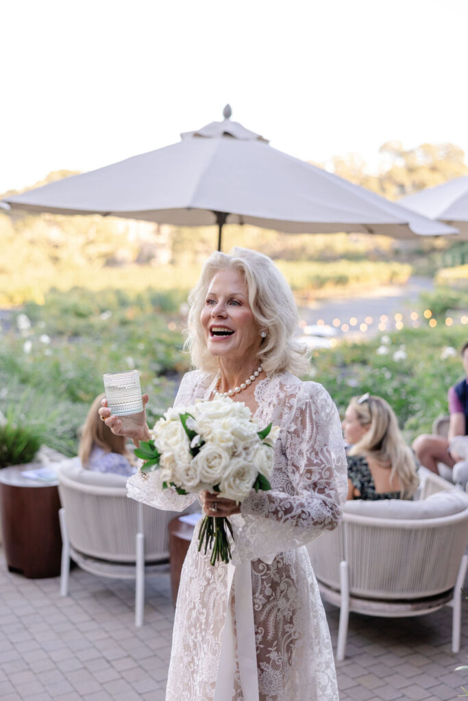 Bride holding a white rose bouquet during cocktail hour at Montage Healdsburg, smiling as guests mingle on the outdoor terrace.