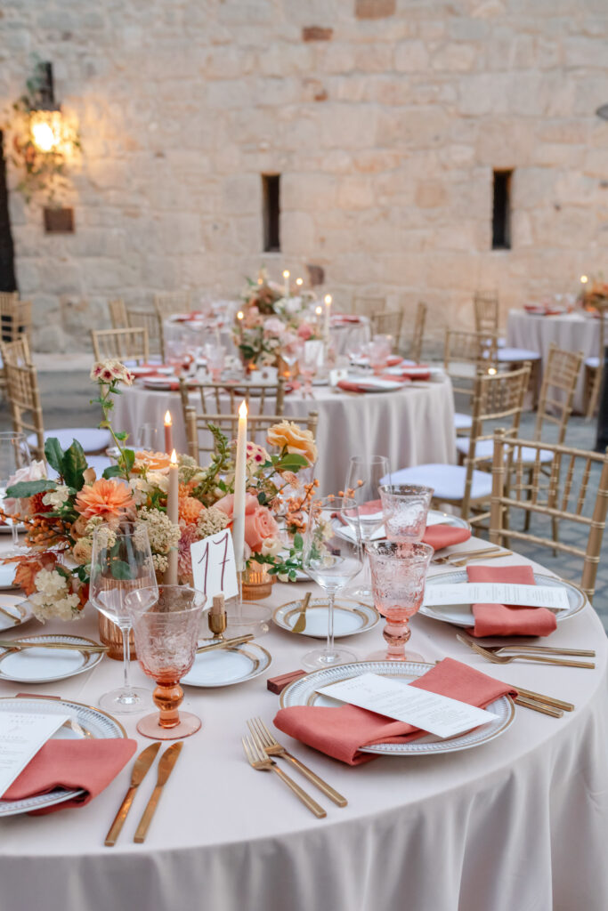 Elegant Napa Valley wedding reception tablescape with gold chiavari chairs, coral linens, and floral centerpieces against a stone winery wall, photographed by Alicia Parks Photography
